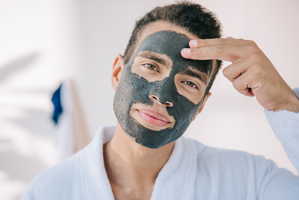 confident young man applying face mask with fingers and looking at camera - Valokuva, kuva