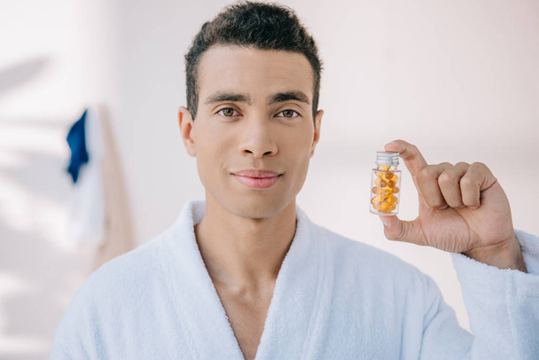 knappe man in badjas Holding fles met vitaminen en kijken naar camera - Foto, afbeelding