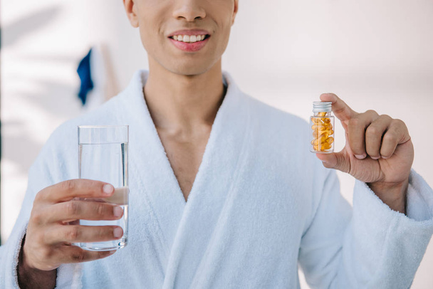 cropped view of man in bathrobe holding bottle with vitamins and glass with water  - Foto, Imagen