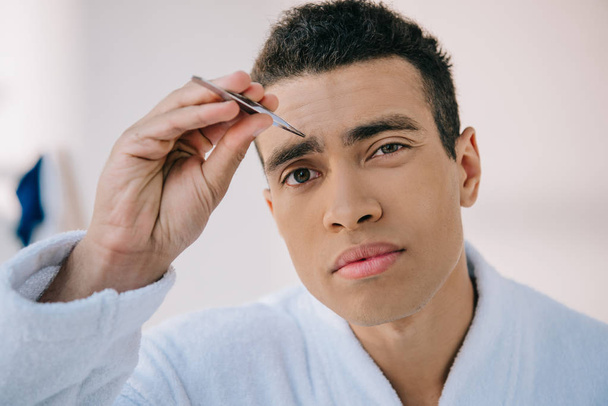 portrait shot of handsome man plucking eyebrows with tweezer and looking at camera - Foto, afbeelding
