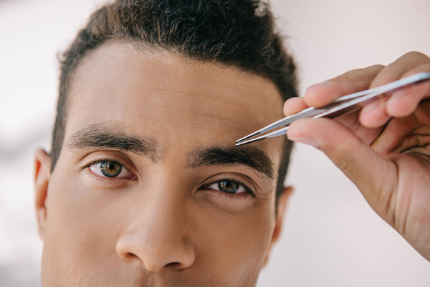 close up view of handsome man plucking eyebrows with tweezer and looking at camera - Photo, Image
