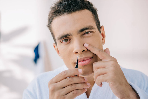 handsome young man plucking nose with tweezer and looking at camera - Φωτογραφία, εικόνα