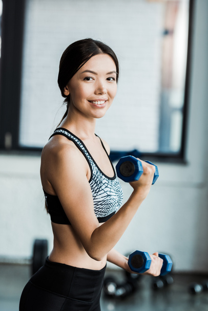 positive and sporty woman holding dumbbells while working out in gym  - Фото, зображення