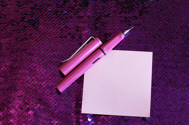 Кусок бумаги для заметок перьевая ручка с кепкой лежат на ярко-розовом фоне ткани с блестками и блестками, свободное место для записей
 - Фото, изображение