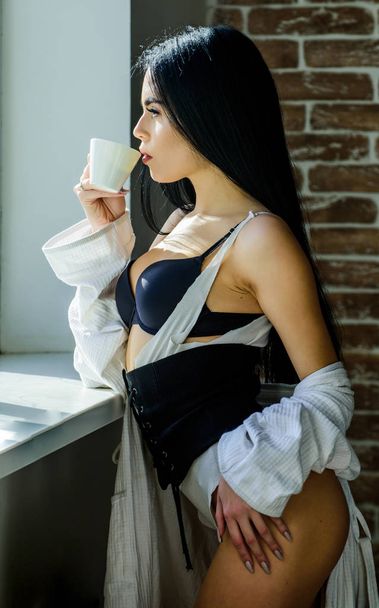 Perfect coffee time. But first coffee. Tranquility and peace. Sip of creamy cappuccino. Morning ritual. Girl sexy breasts drinking coffee near windowsill. Attractive female lingerie relaxing at home - Zdjęcie, obraz