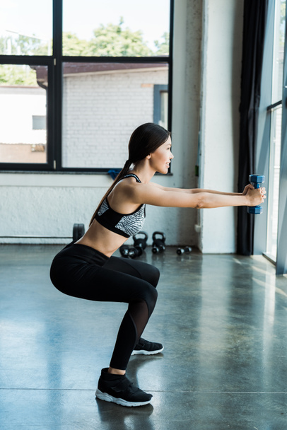 vista lateral de la joven deportista feliz sosteniendo pesas y haciendo ejercicio en cuclillas
  - Foto, imagen