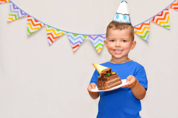A little boy smiles with a dirty mouth in a festive cap and holds a piece of chocolate cake. Celebrates his birthday. - Photo, Image