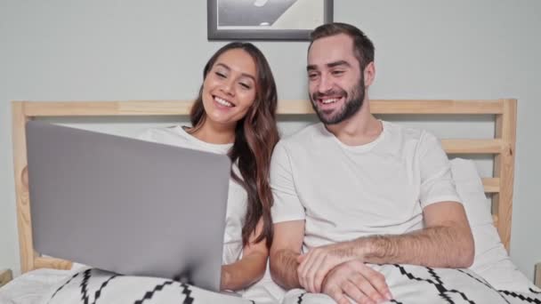 Pleased lovely couple using laptop computer while lying together on bed in bedroom - Footage, Video