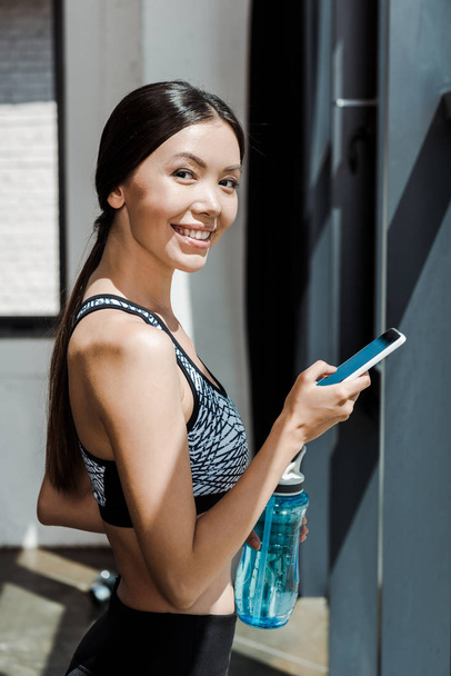 cheerful sportswoman holding smartphone and sport bottle  - Photo, Image