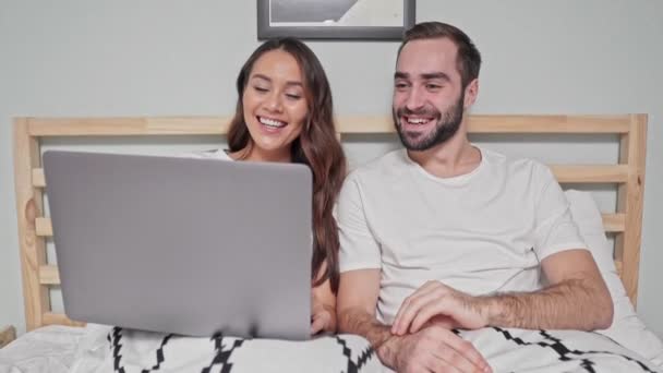 Happy lovely couple talking by videocall on laptop computer while lying together on bed in bedroom - Filmati, video