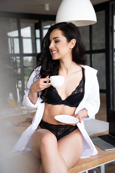 szexi lány fekete fehérnemű és fehér inget iszik kávét csukott szemmel ülve az asztalra a konyhában  - Fotó, kép
