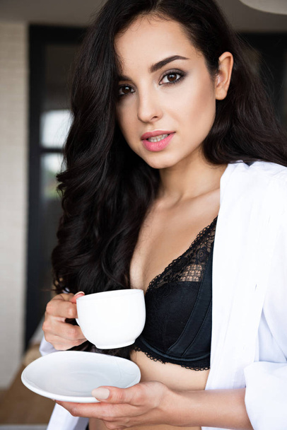 portrait shot of sexy girl in black underwear and white shirt drinking coffee and looking at camera - Fotó, kép