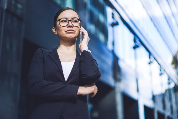Serious business woman dressed in formal wear making concentrated smartphone conversation during break on urban setting, attractive confident female finance professional calling to colleague - Zdjęcie, obraz