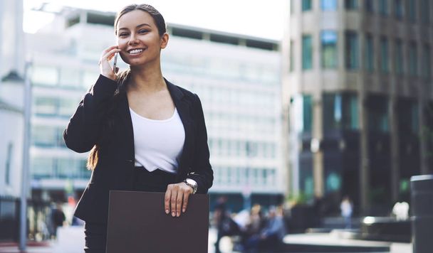 Portrait of cheerful female expert of management looking at camera during positive mobile conversation on publicity area, happy woman making good cellular call via application connected to 4g internet - 写真・画像