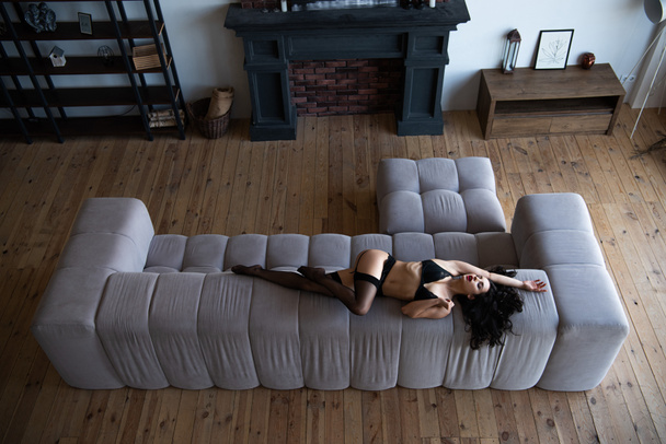 высокий угол зрения сексуальной девушки в черном нижнем белье позирует с закрытыми глазами, лежа на диване в гостиной
  - Фото, изображение
