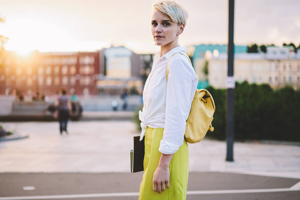 Half length portrait of attractive caucasian hipster girl dressed in stylish outfit and looking at camera during leisure time in evening city, beautiful woman with textbook posing on urban setting - Foto, Imagen