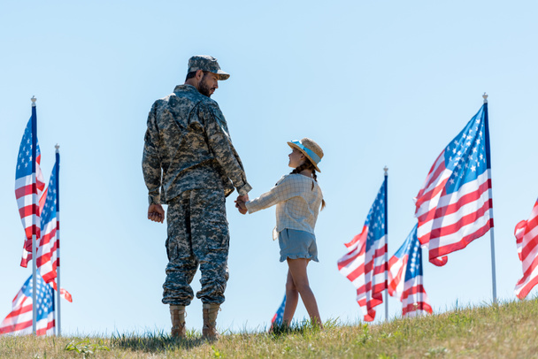 Vater in Militäruniform hält Händchen mit Tochter in der Nähe amerikanischer Flaggen  - Foto, Bild