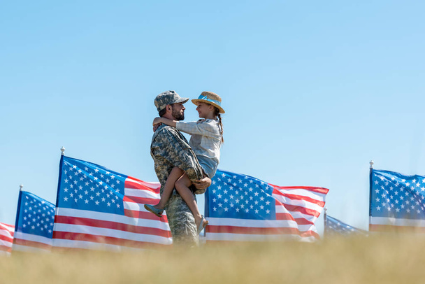 selective focus of military father holding in arms cheerful kid near american flags  - Photo, Image