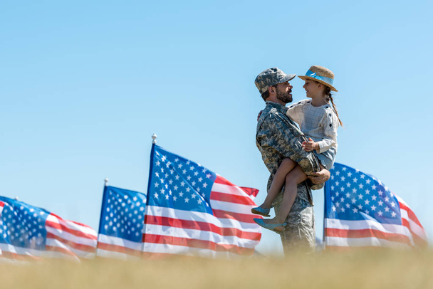 Selektiver Fokus eines Mannes in Militäruniform, der ein fröhliches Kind in der Nähe amerikanischer Flaggen in den Armen hält  - Foto, Bild