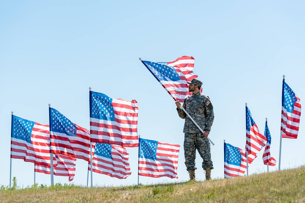 επιλεκτική εστίαση του όμορφου άνδρα σε στρατιωτική στολή και καπάκι στέκεται και κρατώντας αμερικάνικη σημαία  - Φωτογραφία, εικόνα