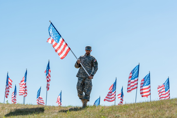 όμορφος στρατιώτης με στρατιωτική στολή και Cap κρατώντας αμερικάνικη σημαία ενώ στέκεται στο γρασίδι  - Φωτογραφία, εικόνα