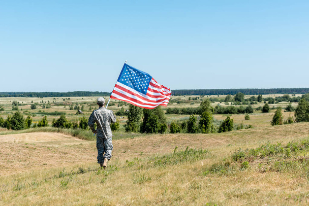 πίσω όψη του στρατιωτικού ανθρώπου που κρατά αμερικάνικη σημαία ενώ περπατάει στο γρασίδι  - Φωτογραφία, εικόνα