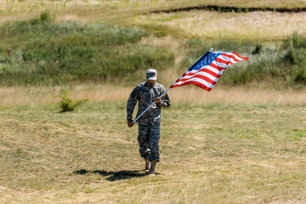 制服と帽子をかぶるハンサムな軍人が夏にアメリカ国旗を掲げて歩く  - 写真・画像