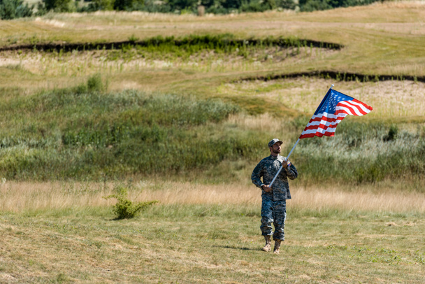 pohledný vojenský muž v uniformní chůzi a drží americkou vlajku v létě  - Fotografie, Obrázek