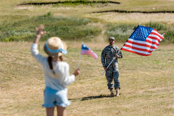 askeri üniformalı adam yakınında Amerikan bayrağı tutarken el sallayarak hasır şapkalı çocuğun arka görünümü  - Fotoğraf, Görsel
