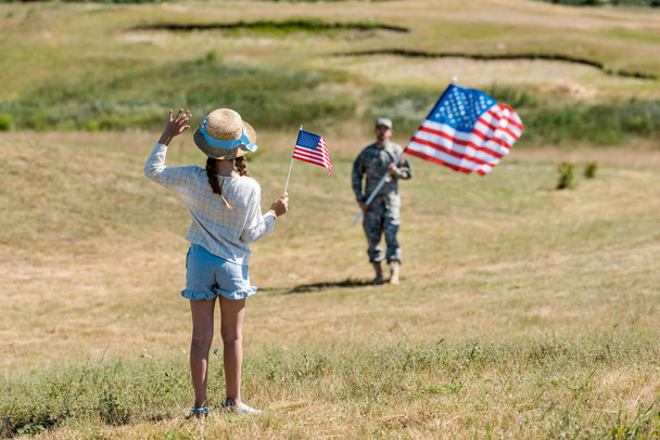 pohled na kluka v slaměném klobouku mávajících rukou a současně držet americkou vlajku poblíž otce ve vojenské uniformě  - Fotografie, Obrázek