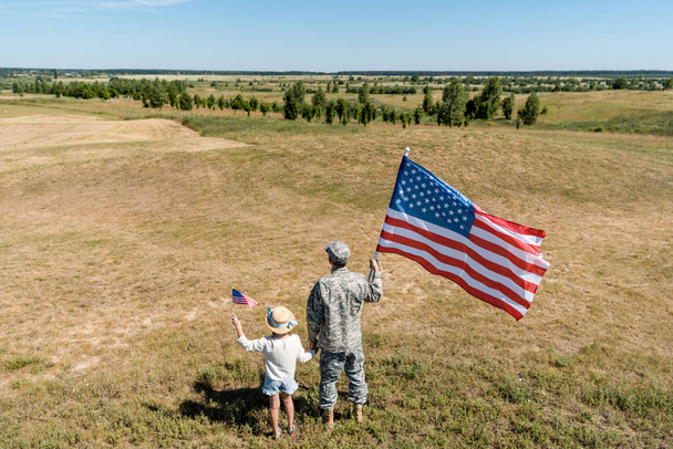 back view of military man and patriotic kid holding american flags  - Фото, зображення