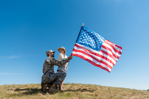 Vater in Militäruniform und glückliches Kind mit amerikanischer Flagge gegen den Himmel  - Foto, Bild