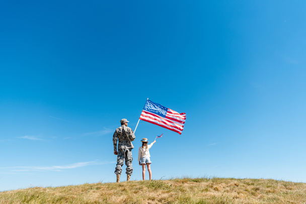 πίσω όψη του παιδιού με ψάθινο καπέλο και στρατιωτικό πατέρα κρατώντας αμερικανικές σημαίες  - Φωτογραφία, εικόνα