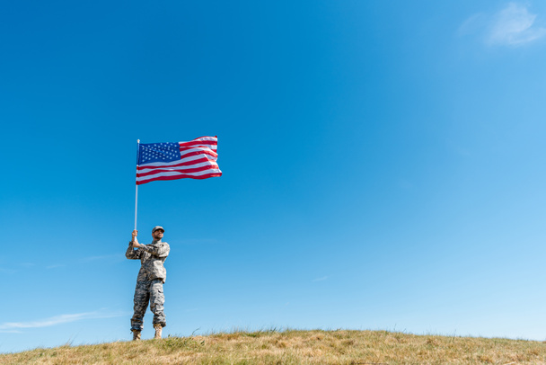 bajo ángulo vista de guapo militar en uniforme caminando con bandera americana con estrellas y rayas
  - Foto, Imagen