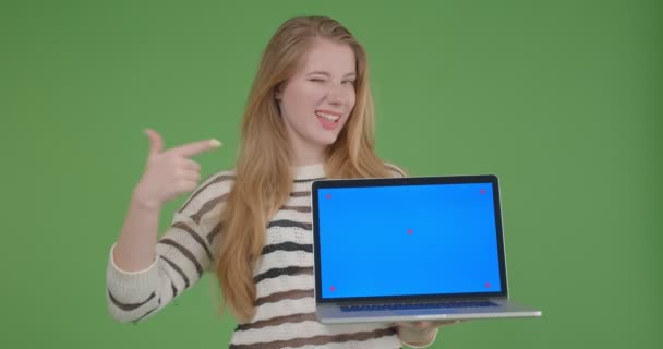Closeup shoot of young pretty caucasian female holding the laptop and showing blue screen to camera smiling cheerfully with background isolated on green - Video