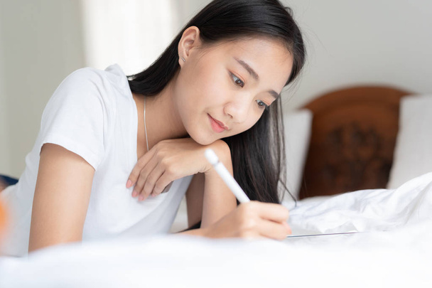 Beautiful Asian woman is drawing on the bed. Asian girl using tablet to learn to draw at her home. - Fotó, kép