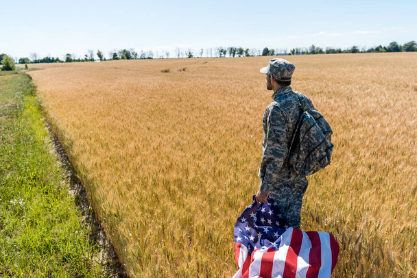 guapo hombre en uniforme militar sosteniendo bandera americana mientras está de pie en el campo
  - Foto, Imagen