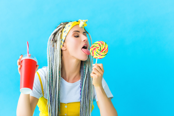 girl with dreadlocks and soda drink licking lollipop isolated on turquoise - Photo, Image