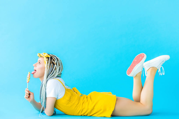 girl with dreadlocks lying and licking lollipop on turquoise - Photo, Image