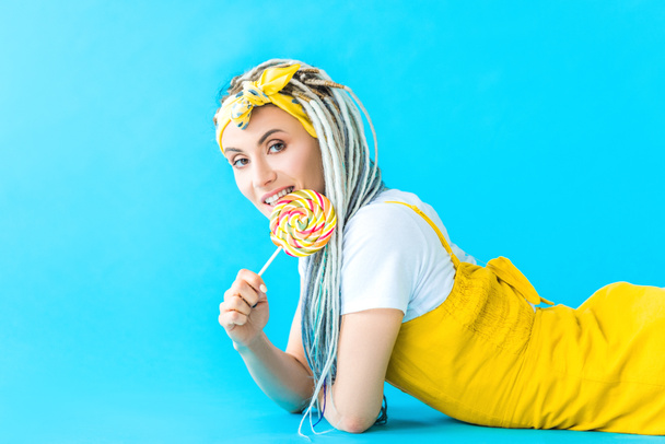 glimlachend meisje met dreadlocks liggend en likken Lollipop op Turquoise - Foto, afbeelding