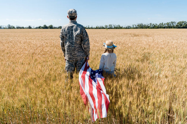 back view of soldier and child standing in field and holding american flag  - Foto, Bild