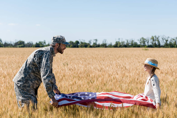soldado en uniforme y niño feliz sosteniendo bandera americana en el campo
  - Foto, Imagen