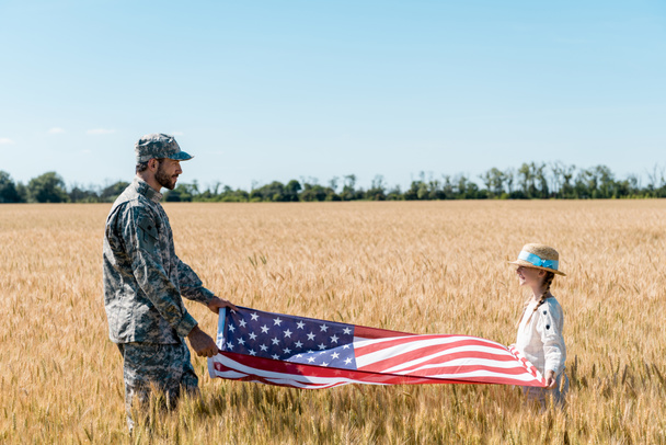 soldier in uniform and cheerful kid holding american flag in field  - Фото, зображення
