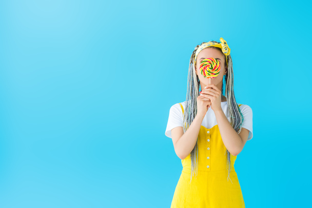 girl with dreadlocks covering face with lollipop isolated on turquoise with copy space - Fotó, kép