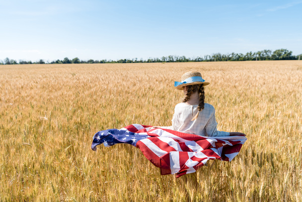back view of kid in straw hat holding american flag in golden field in summertime  - Zdjęcie, obraz
