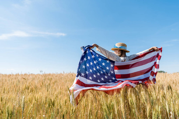 back view of kid in straw hat holding american flag in golden field  - Foto, afbeelding
