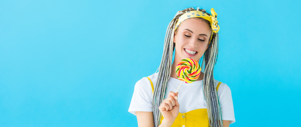 panoramic shot of happy girl with dreadlocks holding lollipop isolated on turquoise - Photo, image