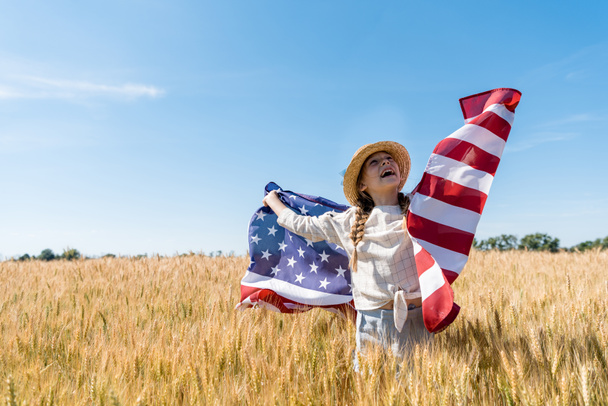cheerful child in straw hat holding american flag in golden field with wheat  - Photo, Image