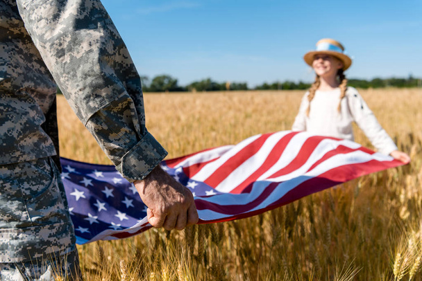 cropped view of man in military uniform holding american flag with daughter in field  - Foto, immagini