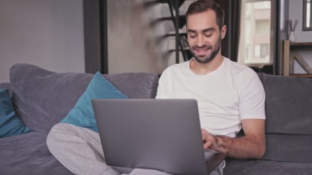 Sorrindo homem barbudo bonito usando computador portátil enquanto sentado no sofá em casa
 - Filmagem, Vídeo